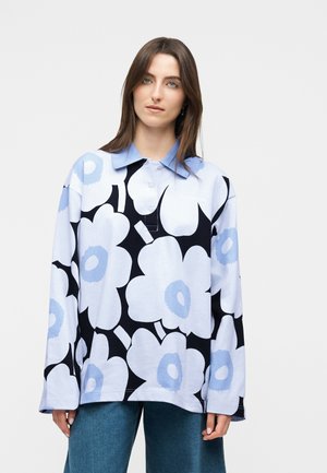 Γυναίκα που φοράει φαρδιά μπλούζα πόλο με μακριά μανίκια και μεγάλα σχέδια με λευκά και ανοιχτό μπλε λουλούδια, καθώς και μπλε τζιν, στέκεται μπροστά σε μονόχρωμο φόντο.