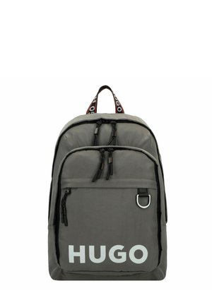 Mochila de nailon gris con tiradores de cremallera negros y un bolsillo frontal. Gran logo blanco de "HUGO" y correa contrastante con marca.