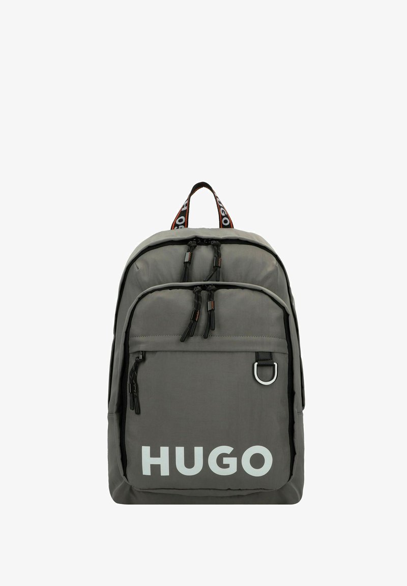 Mochila de nailon gris con tiradores de cremallera negros y un bolsillo frontal. Gran logo blanco de "HUGO" y correa contrastante con marca.
