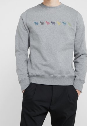 Grauer Pullover mit sieben kleinen bunten Zebras, die quer über die Brust gestickt sind, getragen zu schwarzen Hosen, Hände in den Taschen.