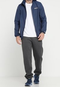 Marinblå vindjacka med huva, framdragkedja och logopatch. Tillsammans med grå sweatpants och marinblå sportskor. Vit t-shirt under.