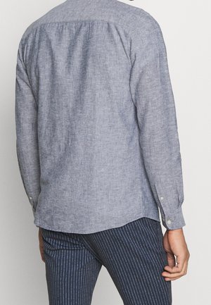 Homme portant une chemise bleu clair à manches longues et un pantalon bleu foncé à fines rayures, tourné de dos, main détendue le long du corps.