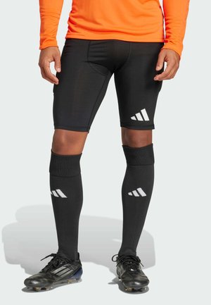 Black athletic shorts with a tight fit, featuring a white Adidas logo. Paired with black knee-high socks and black soccer cleats.