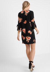 Woman walking away wearing black dress with pink floral print and three-quarter sleeves, paired with black high-heeled ankle-strap shoes.