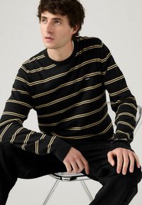 Un giovane seduto su una sedia, vestito con un maglione nero con sottili righe orizzontali gialle e pantaloni neri, che guarda leggermente in alto a sinistra.