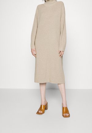 Beiges, langärmliges, geripptes Midikleid mit Stehkragen, kombiniert mit hellbraunen, offenen Blockabsatz-Sandalen an einer stehenden Person.