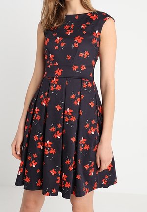 Ærmeløs sort kjole med rødt blomsterprint, tætsiddende talje og udvidet skørt båret af en person, der står mod en ensfarvet baggrund.