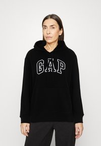 Svart hoodie med en stor grå "GAP"-logotyp på framsidan, med snörning i huvan och kängurufickor. Mjuk tyg med ribbade manschetter.
