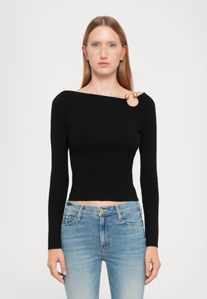 Black ribbed long-sleeve top with an asymmetrical neckline, featuring a circular gold accent and a fitted waist, paired with blue denim jeans.