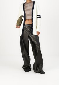 Weißer Cardigan mit dunkelblauen Streifen, geknöpft, über einem braun gestreiften Crop-Top. Schwarze, weit geschnittene Lederhosen und eine grüne Handtasche.