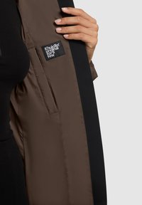 Eine Hand hält eine geöffnete schwarz-braune Jacke und zeigt auf die Innentasche mit einem schwarzen Etikett, auf dem in weißem Text "Khujo original for a few" steht.