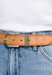 Hellbrauner Wildledergürtel mit silberner Schnalle, der um die Taille einer blauen Jeans getragen wird, mit einem eingesteckten weißen Hemd.