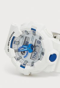 Montre-bracelet G-Shock blanche avec des accents bleus, cadran rond, affichages numérique et analogique, boutons texturés et un design robuste et résistant aux chocs.