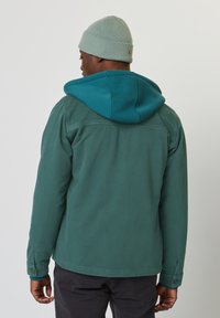 Türkise Baumwolljacke mit einem blauen Kapuzenpullover darunter, mit Knopfmanschetten und schlichter Gestaltung. Kombiniert mit einer hellgrünen Strickmütze.
