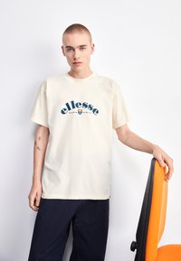 Camiseta corta de manga corta color crema con un logo gráfico azul y naranja. El tejido parece suave y el corte es holgado, complementado con pantalones oscuros.