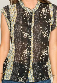 Blouse noire à boutons avec un motif floral aux accents jaunes et blancs. Les manches sont de style capuchon et le col est volanté.