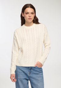 Sweter z dzianiny w kolorze ecru, z okrągłym dekoltem i ściągaczami przy rękawach. Połączony z jasnoniebieskimi dżinsami dla uzyskania swobodnego wyglądu.