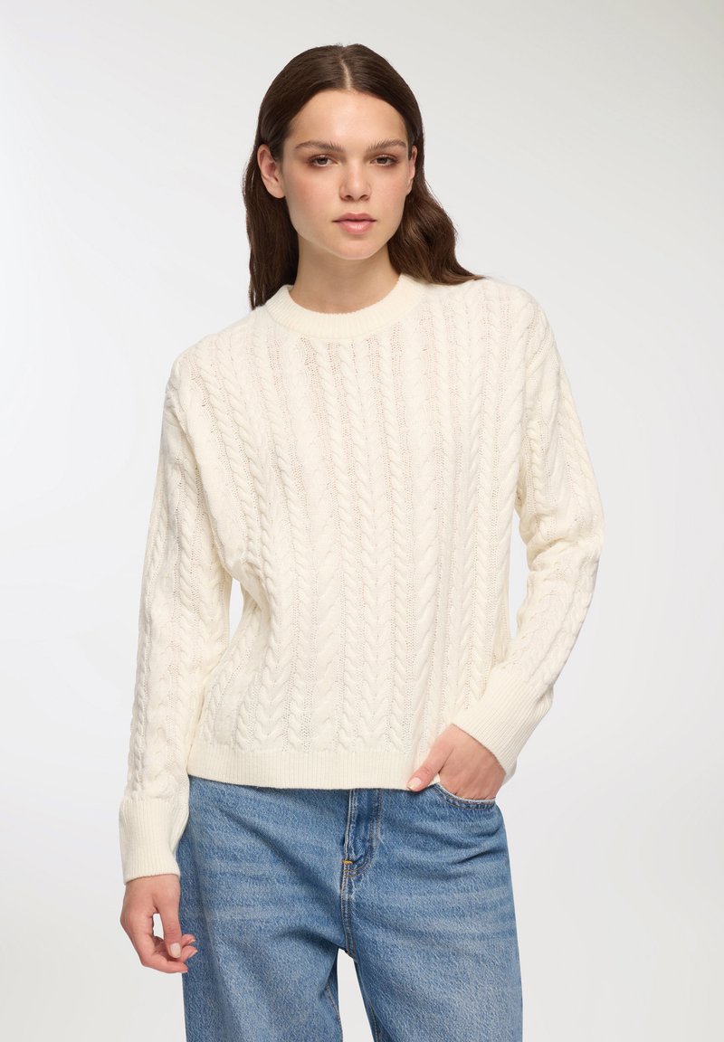 Sweter z dzianiny w kolorze ecru, z okrągłym dekoltem i ściągaczami przy rękawach. Połączony z jasnoniebieskimi dżinsami dla uzyskania swobodnego wyglądu.