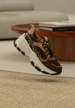 Sneaker chunky con dettagli beige, marroni e stampa leopardata che fluttano sopra un tappeto beige, con un tavolo di legno e libri sullo sfondo.