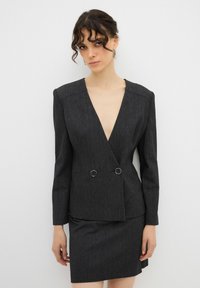 Zwarte, geweven blazer met een diepe V-hals, dubbele ringdetails en gestructureerde schouders, gecombineerd met een bijpassende aansluitende rok.