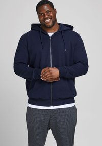 Navyfarbene Zip-Hoodie mit Kapuze, Fronttaschen, gerippten Bündchen und lockerer Passform, getragen über einem weißen Shirt und gemusterten Hosen.