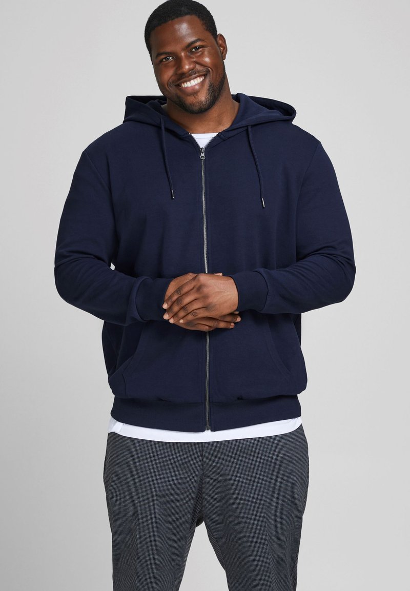 Navyfarbene Zip-Hoodie mit Kapuze, Fronttaschen, gerippten Bündchen und lockerer Passform, getragen über einem weißen Shirt und gemusterten Hosen.