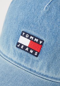 Lyseblå denimkasket med et broderet rektangulært logo-patch i marineblå, hvid og rød med teksten "TOMMY JEANS." Har en buet skygge.