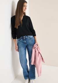 Jeune femme aux cheveux longs et bruns, portant un haut noir et un jean évasé, tenant une veste rose, appuyée contre un mur blanc.