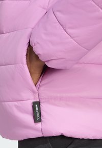 Růžová péřová bunda s rukou sahající do boční kapsy. Štítek zní "CLIMA WARM", což zdůrazňuje tepelnými vlastnosti bundy.