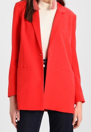 Femme portant un blazer rouge vif sur un col roulé blanc côtelé et un pantalon foncé, avec des boucles d’oreilles à pampilles roses visibles.