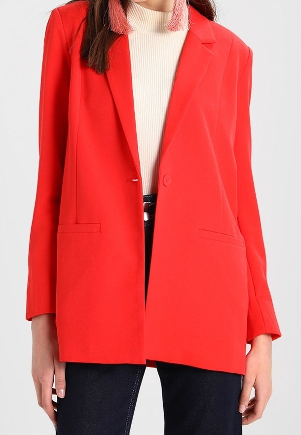 Femme portant un blazer rouge vif sur un col roulé blanc côtelé et un pantalon foncé, avec des boucles d’oreilles à pampilles roses visibles.