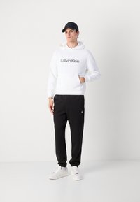 Hanorac alb cu glugă și logo-ul negru "Calvin Klein", asortat cu pantaloni sport negri cu un logo brodat și adidași albi. Poartă o șapcă neagră.