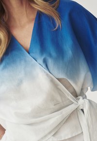 Top envolvente en ombré de azul a blanco con mangas cortas. Confeccionado en una tela ligera, presenta un cierre de lazo y una textura suave.