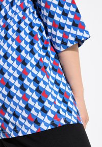 Camicia a maniche corte in cotone blu, caratterizzata da un motivo geometrico di triangoli in bianco, nero, rosso e navy. Dettaglio di orlo svasato.