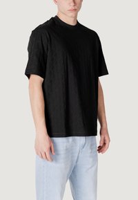 Maglietta nera a maniche corte con un motivo geometrico testurizzato, scollo rotondo e vestibilità ampia. Abbinata a jeans azzurri chiari.