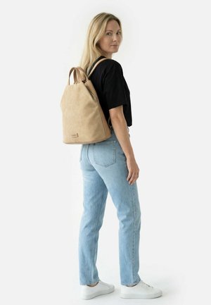 Žena v černém tričku a světlých modrých džínách s béžovým batohem stojí bokem a ohlíží se přes rameno na bílém pozadí.