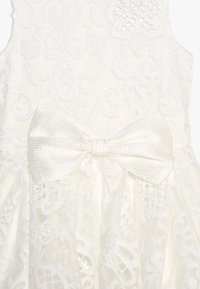 Witte kanten jurk met ingewikkelde bloemenpatronen, een gestructureerde stof en een grote geweven strikaccent rond de taille.