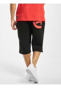 Svarte shorts til kneet med en rød sirkulær logo-lomme detalj, parret med hvite joggesko. Rød skjorte er synlig i kanten bak.