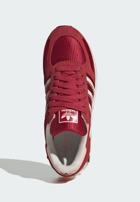 Κόκκινο αθλητικό παπούτσι με υφή στο επάνω μέρος, υλικά από ύφασμα και σουέτ, τρεις λευκές ρίγες και επενδεδυμένη γλώσσα με το λογότυπο της Adidas.