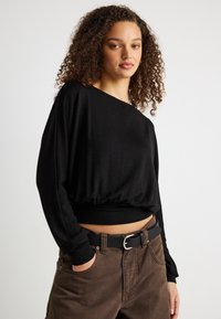 Νεαρή γυναίκα με σγουρά μαλλιά, φορώντας μαύρο μακρυμάνικο τοπ και καφέ παντελόνι με μαύρη ζώνη, στέκεται με το ένα χέρι στην τσέπη.