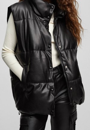 Femme portant une doudoune matelassée en cuir noir sur un pull côtelé crème et un pantalon cargo en cuir noir, les mains dans les poches de la doudoune, cheveux bouclés mi-longs.