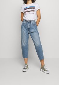 Bílá tričko s grafickým designem a krátkými rukávy, spárované s světle modrými zkrácenými džíny s neomítnutým lemem a zelenými teniskami.