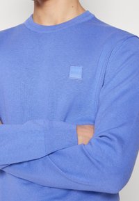 Pull en tricot bleu clair avec un col rond, présentant un petit patch logo carré sur la poitrine. Le tissu a une texture lisse et des poignets côtelés.