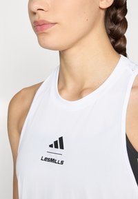 Tricou sportiv fără mâneci, alb, cu detalii în negru pentru logo-urile "LesMills" și adidas. Textură netedă, decolteu rotund și margine ușor curbată.