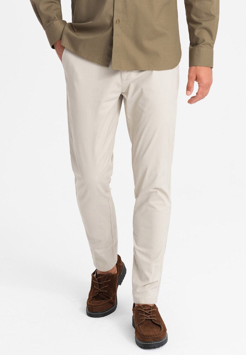 Pantalones elásticos beiges de corte slim, con una textura suave y pierna ajustada, combinados con zapatos de ante marrones.