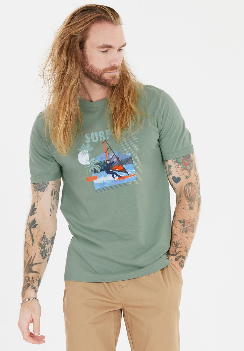 Vihreä lyhythihainen t-paita, jossa on värikäs surffausgrafiikka, kevyt kangas, pyöreä kaula-aukko ja rento istuvuus, yhdistettynä vaaleisiin housuihin.