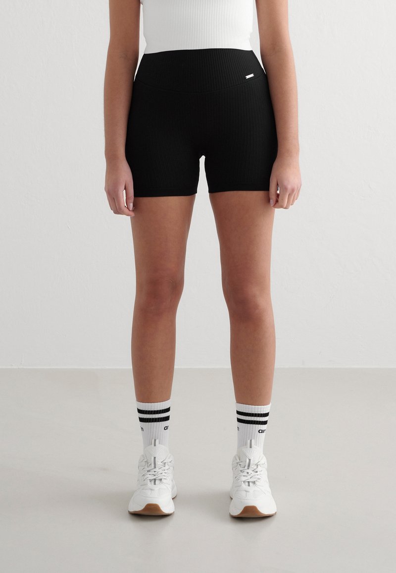 Sorte ribbede shorts med høy midje, parret med hvite joggesko og stripete sokker. Den glatte teksturen og tettsittende formen fremhever komfort.