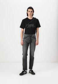 Zwart t-shirt met grafisch ontwerp, gecombineerd met donkergrijze jeans en zwarte sneakers met witte strepen. Model staat tegen een effen achtergrond.