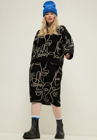 Schwarzes Strickensemble mit abstrakten Gesichtsmustern in Beige. Beinhaltet einen lockeren Pullover und einen taillierten Rock, kombiniert mit schwarzen Plateau-Stiefeln und einer blauen Mütze.