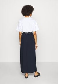 T-shirt blanc à manches courtes associé à une longue jupe bleu marine avec une taille élastique portant un logo. Chaussures noires à enfiler visibles.